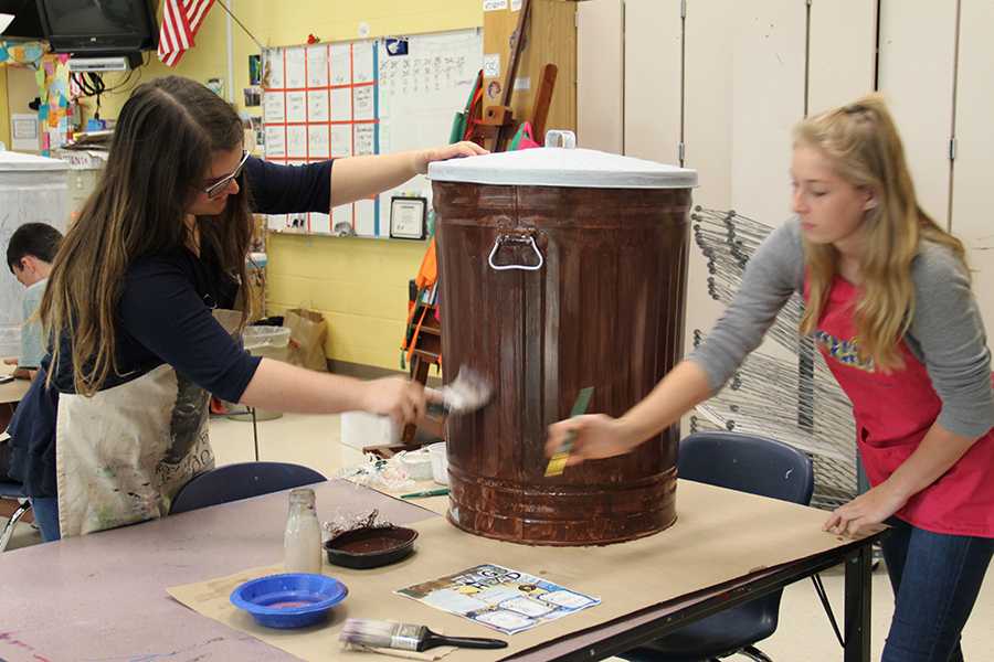 Art students Lorna Cameron (left) and Cassidy Joyce work on the OB Cans project in art class last May. Students will be painting more cans for this summer at Artrageous on Saturday. PHOTO BY AMY WRENN/NIGHTHAWK NEWS